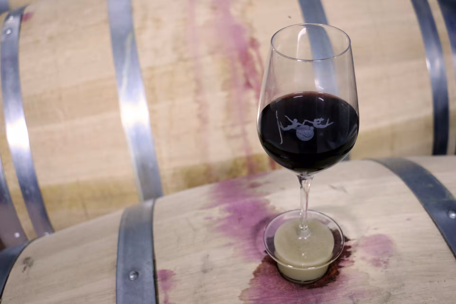 Branded red wine glass resting on a rustic oak barrel during a free-flow guided wine tasting tour at a winery