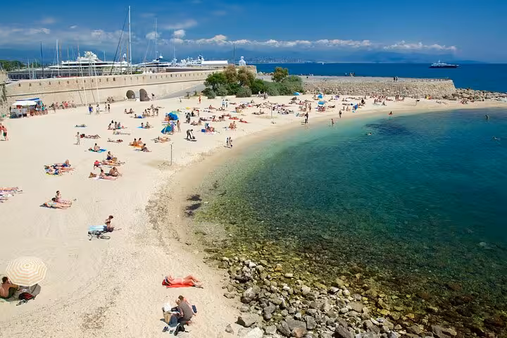 Sunny French Riviera beach with turquoise water, sunbathers on white sand, and yachts in harbor beyond stone wall