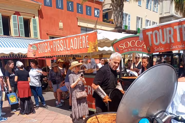 Bustling French Riviera street market with socca stalls and people enjoying local cuisine under a clear blue sky.