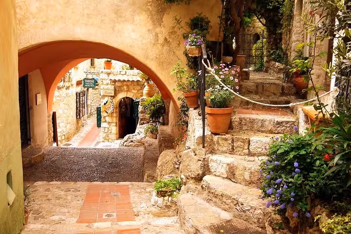 Charming stone alleyway with arched passageway, terracotta pots, flowering plants, and rustic steps in French Riviera village