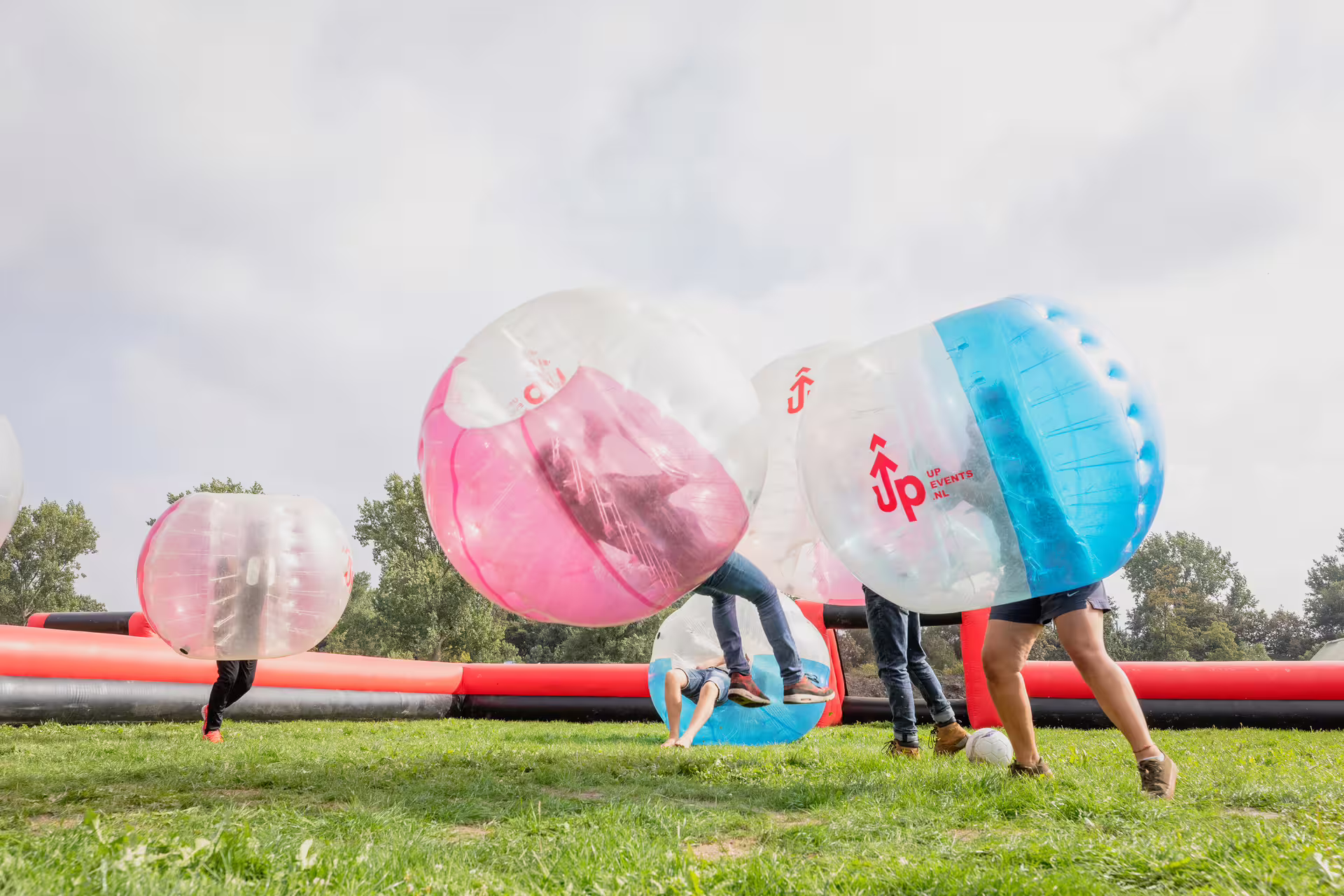 Friends play bubble football in Amsterdam, bumping in inflatable zorbs on a grassy outdoor pitch