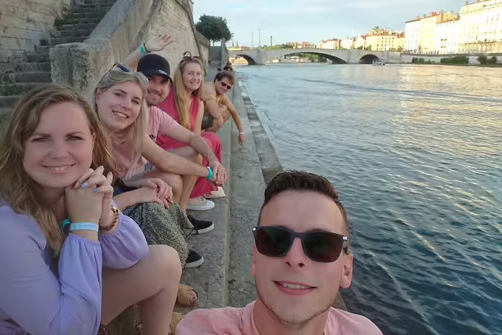 Group of friends sitting along Lyon riverbank during guided walking tour on sunny day