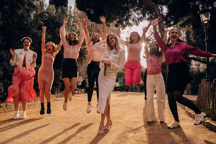 Friends jump mid-air on a sunlit Barcelona park path as a guest in a white dress holds flowers during a lively photoshoot.