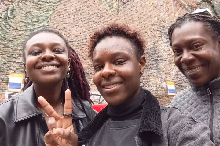 Three smiling women posing for a selfie in front of a brick wall with street art in Manchester