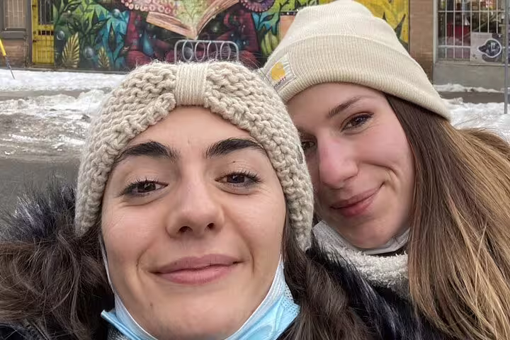 Two smiling women wearing winter hats pose for selfie in front of colorful street art mural in Ottawa