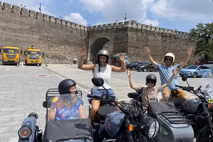 Group of friends raise arms in excitement during a sidecar tour, parked near ancient Beijing city gate.