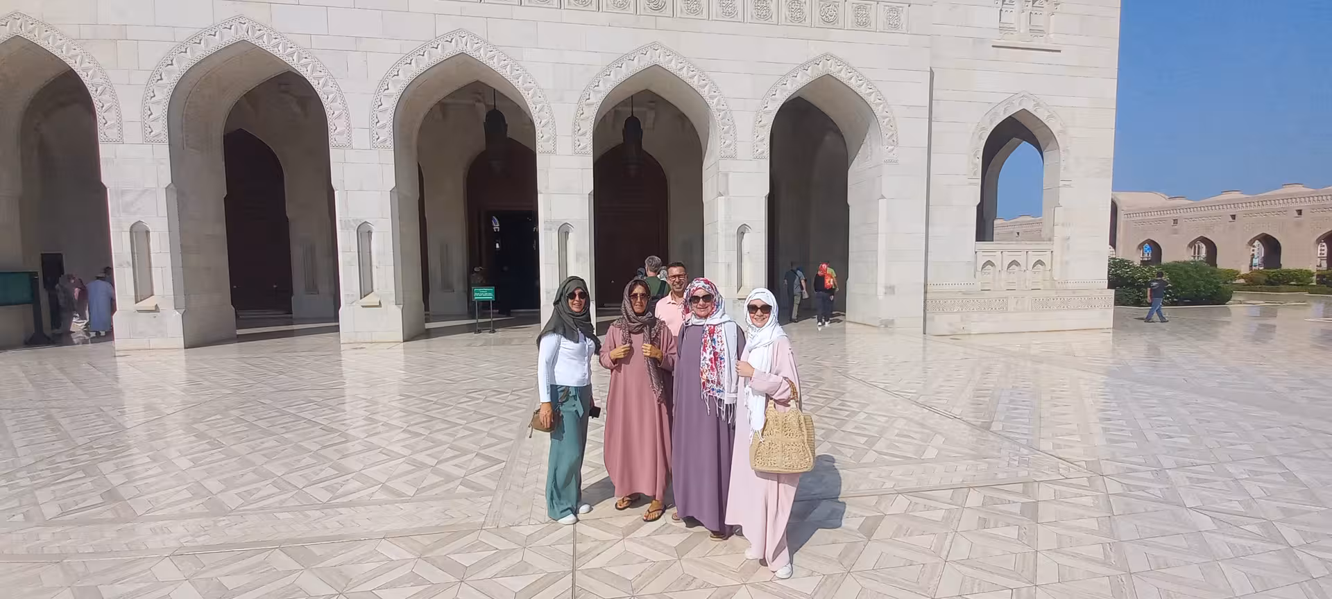 Friends in modest dress exploring the marble courtyard and arches of Sultan Qaboos Grand Mosque on a Muscat city tour