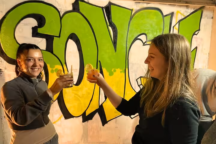 Two friends toast cocktails in front of a bright graffiti wall on the Sip and Spray with Locals tour