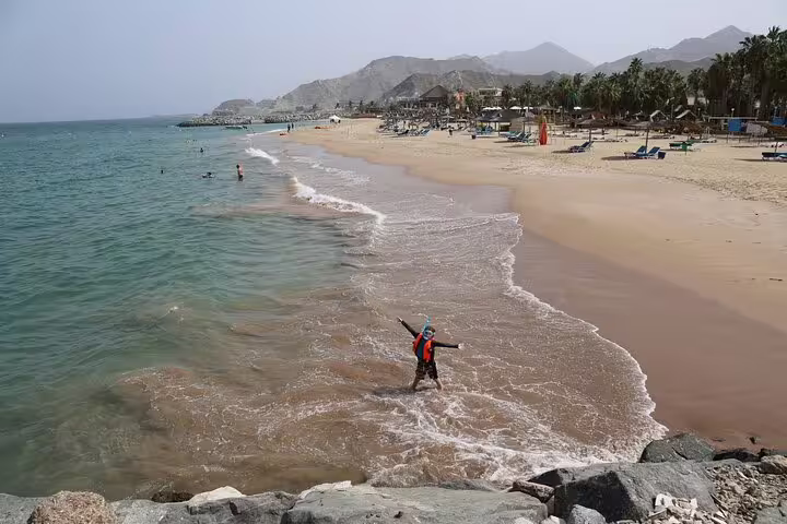 Tourists enjoying the sandy beaches and clear waters of Fujairah, surrounded by mountains and palm trees.