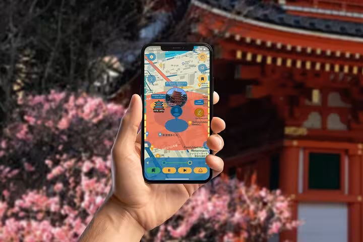 Hand holding phone displaying self-guided GPS tour map near vibrant red Fukuoka temple with cherry blossoms