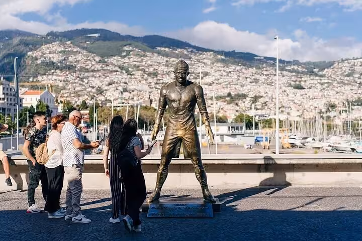 Visitors admire a prominent statue in Funchal with scenic hillside and marina views in the background.