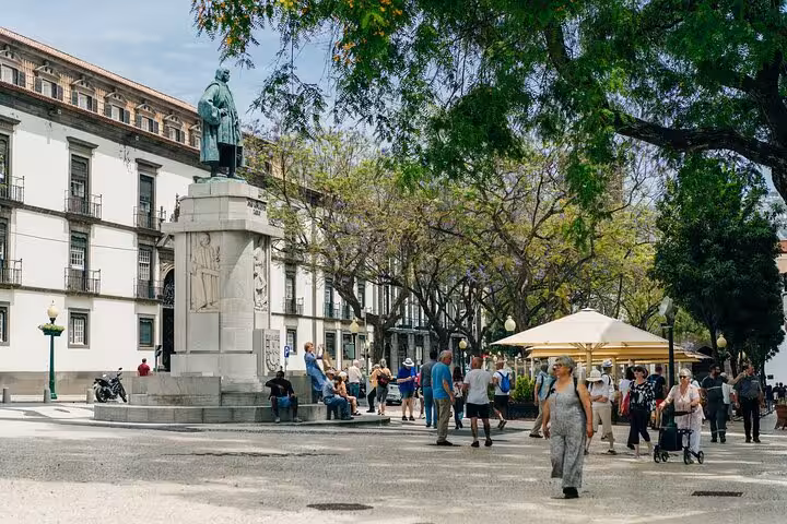 Lively Funchal square with historical statue, locals, and tourists enjoying a guided city walk under lush trees.