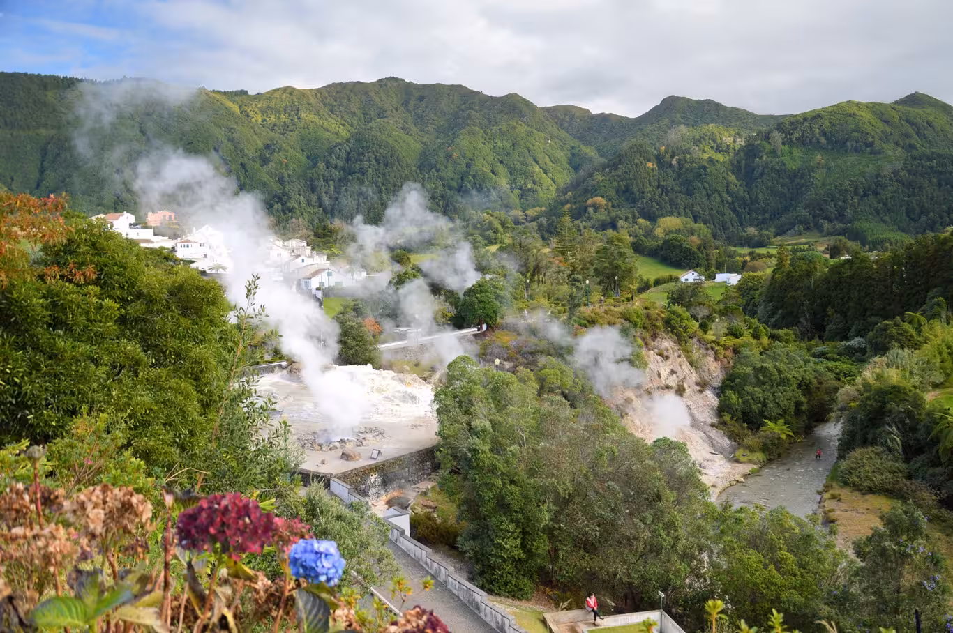 Steam rises from Furnas geothermal valley surrounded by lush green mountains and canyoning river on São Miguel, Azores