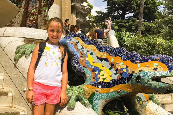 A child smiles next to the colorful mosaic salamander at Park Güell, a highlight of the 2-day Gaudí, Sagrada Familia, and Montserrat tour.