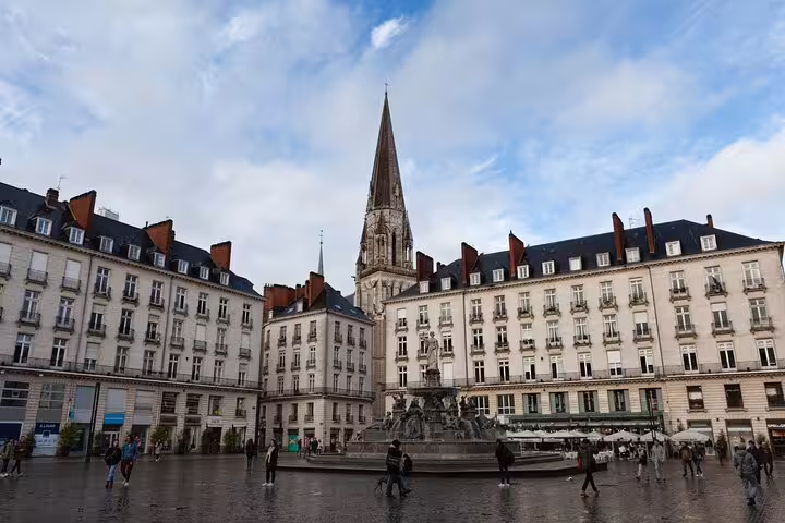 Historic city square with ornate fountain, Haussmann buildings, and Gothic church spire under cloudy sky