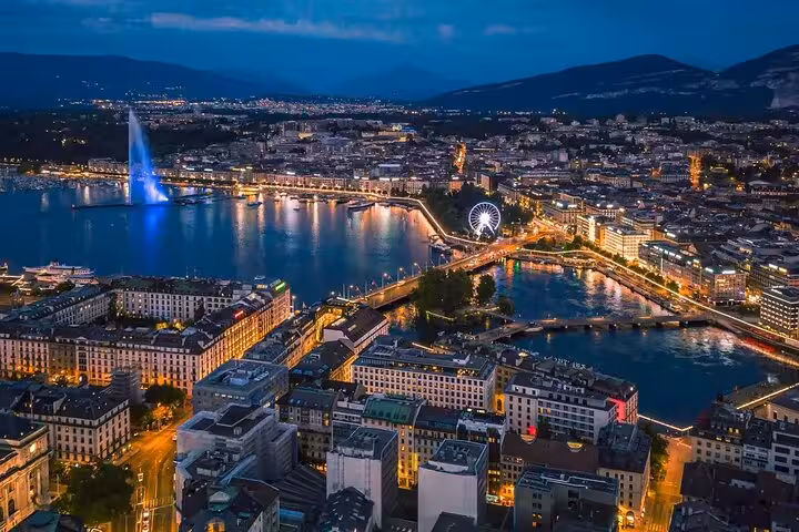 Aerial view of Geneva at night showcasing the illuminated cityscape and Jet d'Eau fountain along the waterfront.
