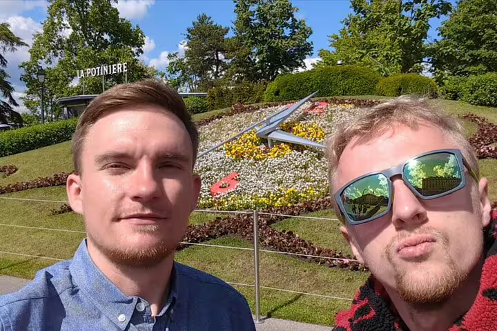 Two men taking a selfie in front of La Potinière flower clock in Geneva on a sunny day
