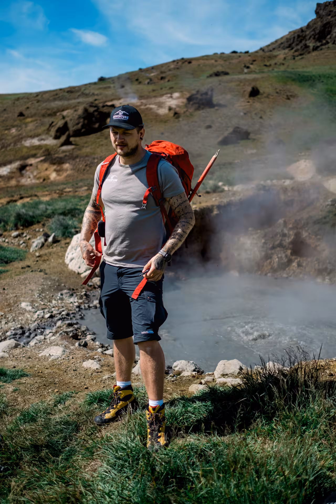 Tattooed hiker with red backpack walking near steaming geothermal hot spring in Iceland volcanic valley