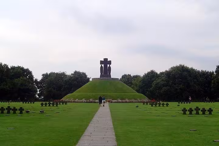 German military cemetery with dark cross grave markers and central memorial sculpture on grassy mound, Normandy