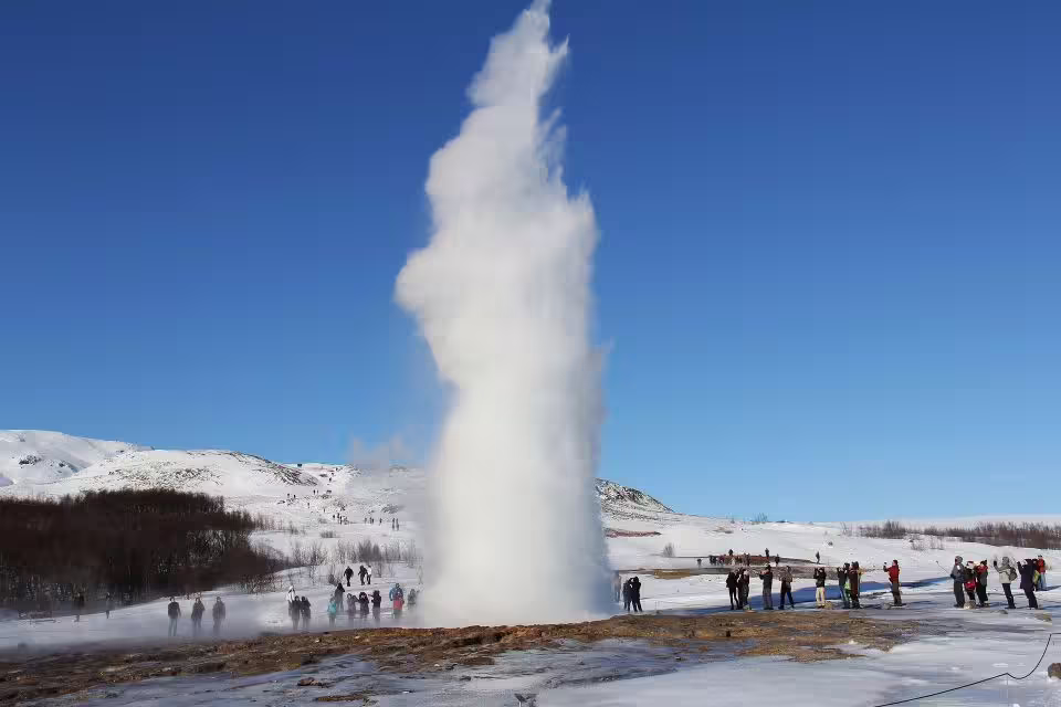 Geyser erupts against a clear blue sky in Iceland's Golden Circle, surrounded by snow and tourists capturing the moment.
