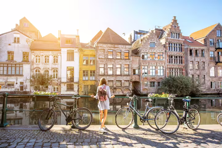 Woman with backpack enjoys canal view in Ghent, Belgium, showcasing charming historic architecture on a sunny day.