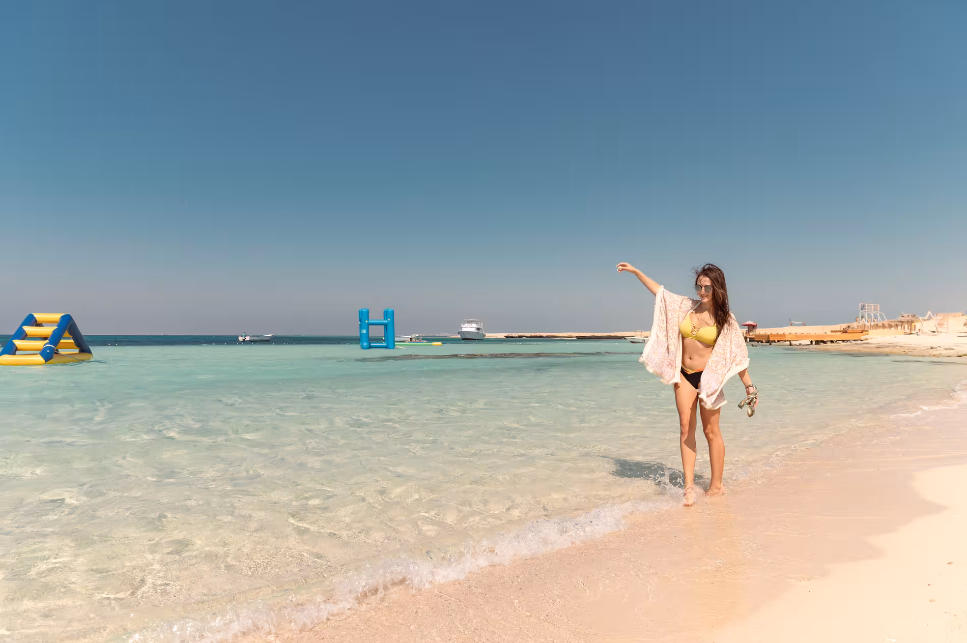 Woman in yellow bikini walking along clear turquoise shoreline at Giftun Island with inflatable water park