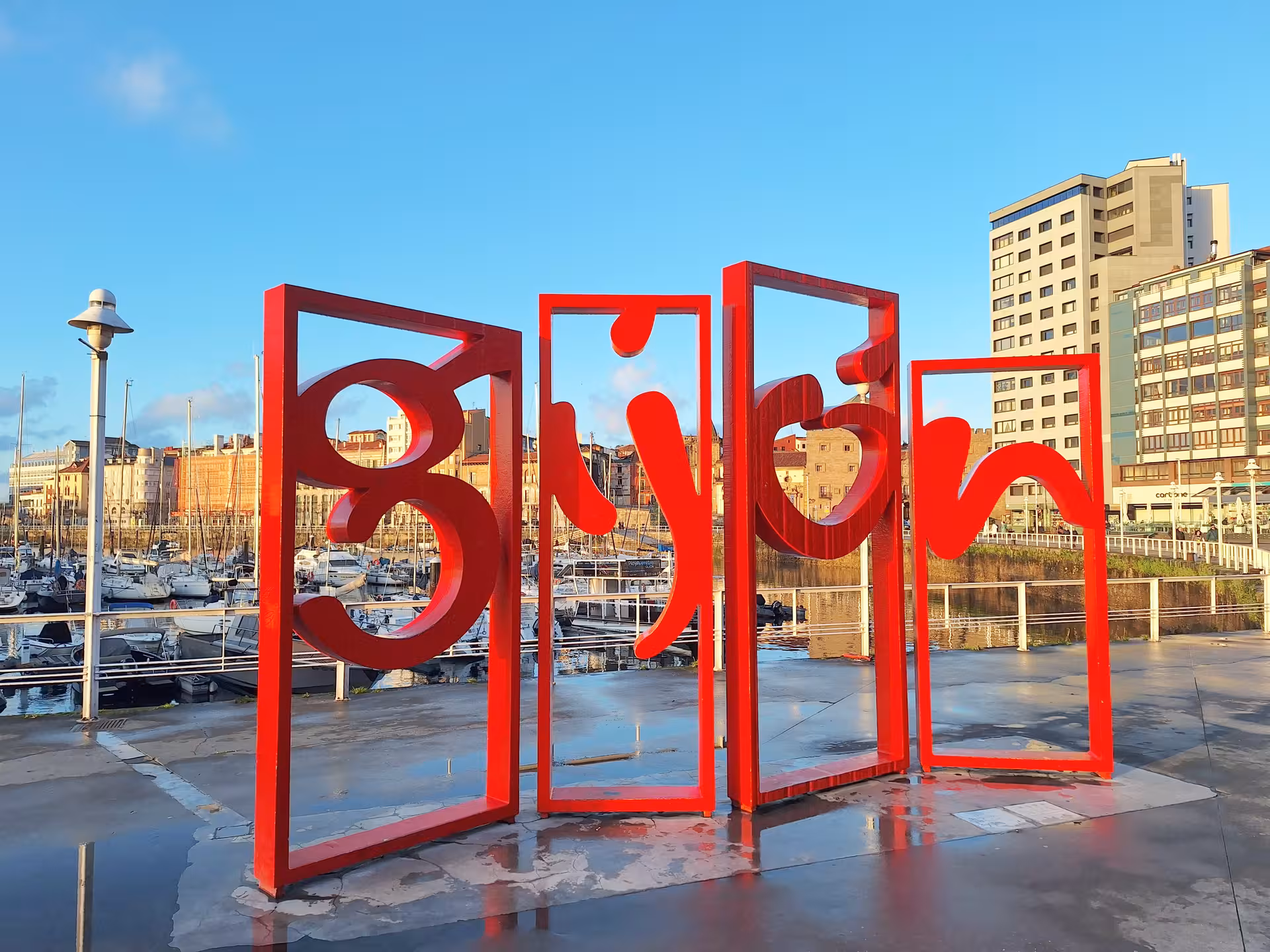 Vibrant red Gijon sign against a backdrop of the cityscape and marina, highlighting the city's iconic waterfront.