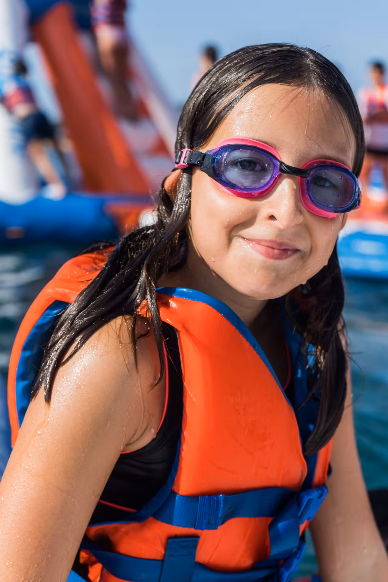 A smiling girl wearing pink goggles and a life vest enjoys a fun day at an inflatable waterpark on the water.