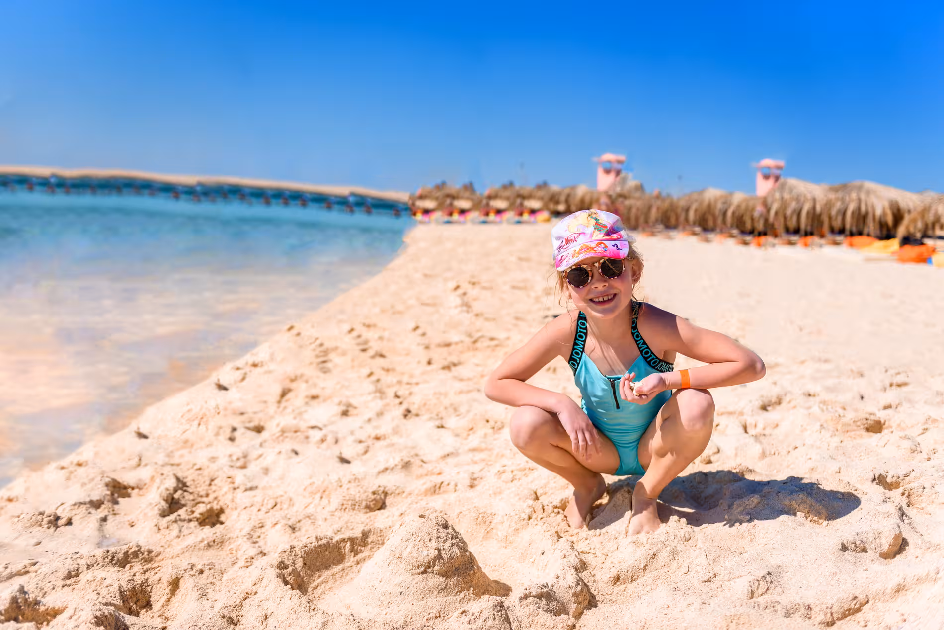 Young girl in teal swimsuit crouching on white sand beach at Giftun Island on a sunny day