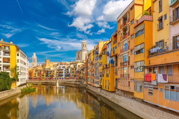 Colorful riverside buildings of Girona reflect in the Onyar River under a vibrant blue sky, perfect for a Costa Brava tour.