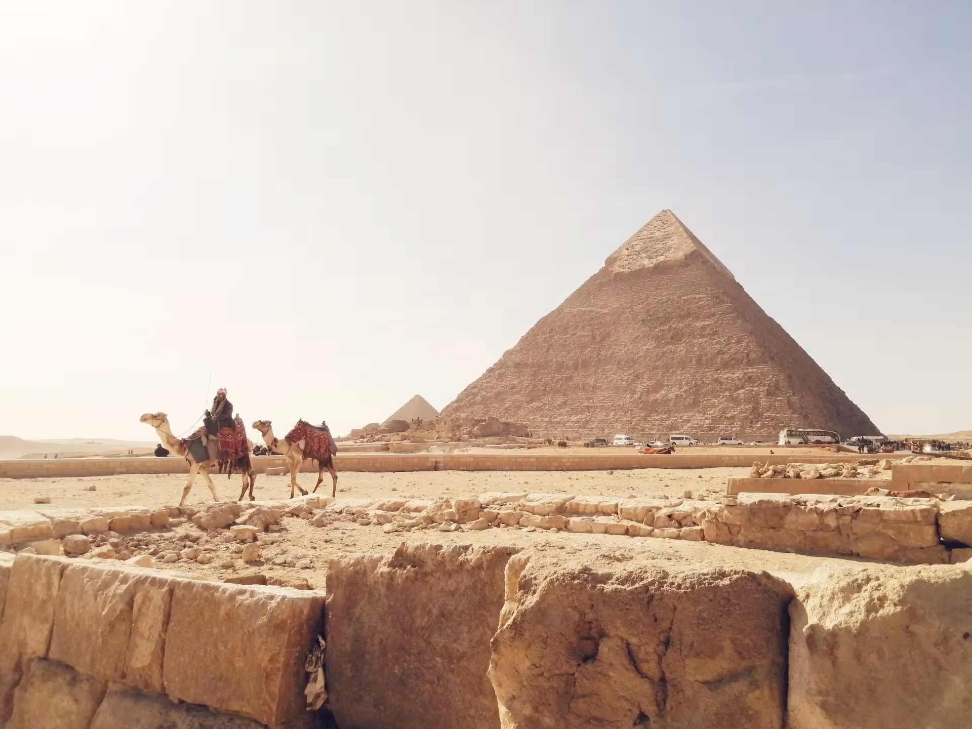 Camel rider passing ancient ruins with the Great Pyramid of Khafre rising against a hazy sky at Giza