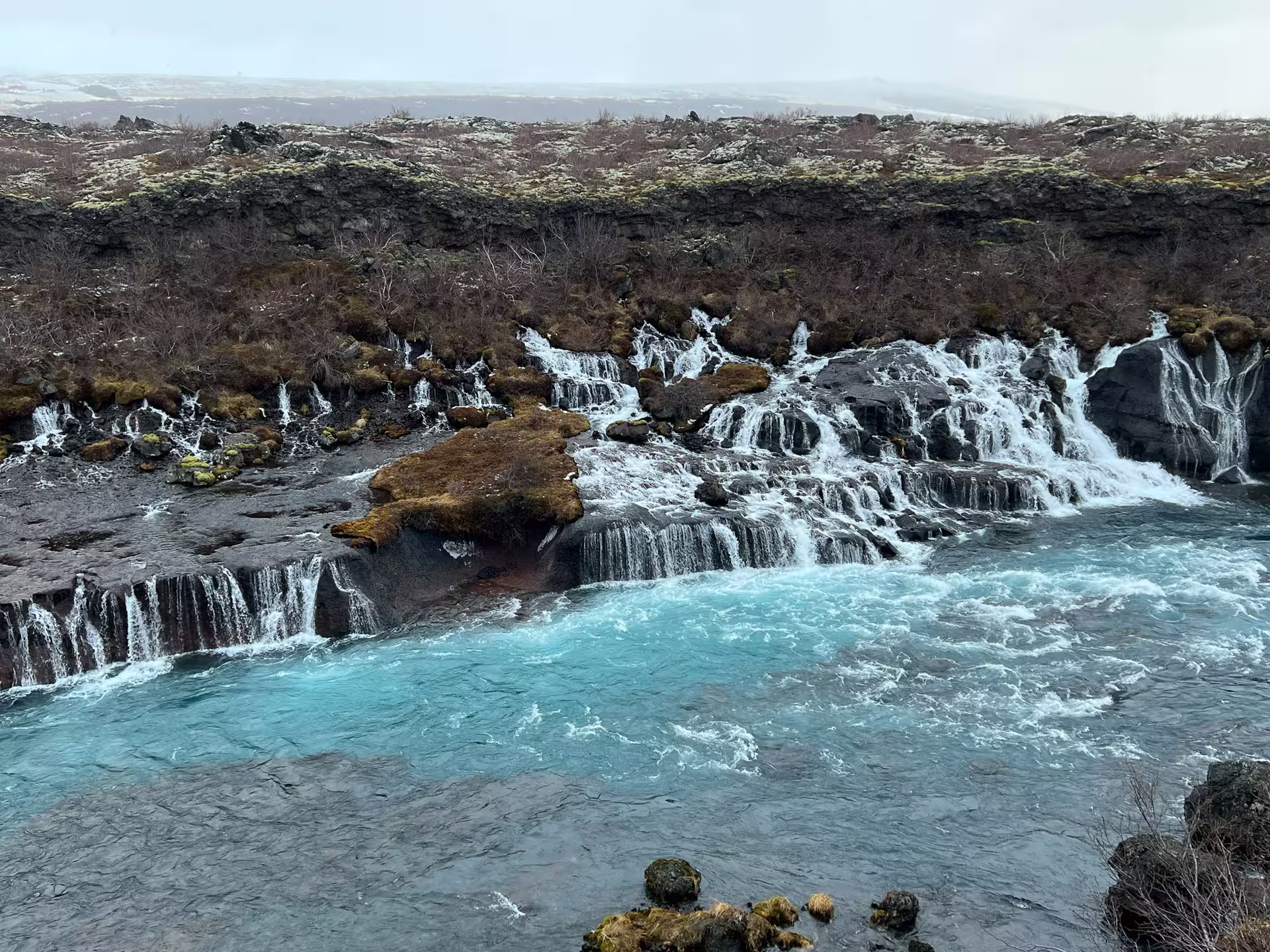 Turquoise glacial river with cascading waterfalls flowing over dark lava rock formations in Iceland