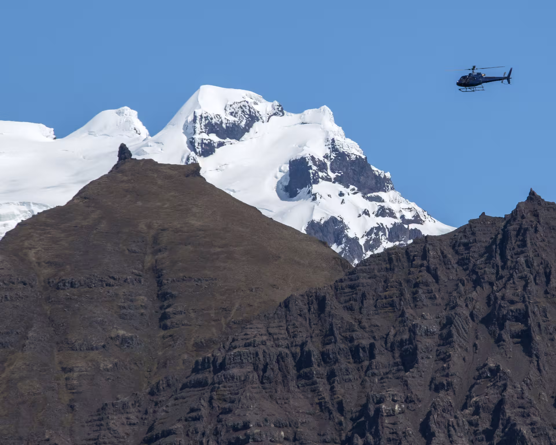 Helicopter flying over snow-capped glacial mountain peaks in Iceland under clear blue sky