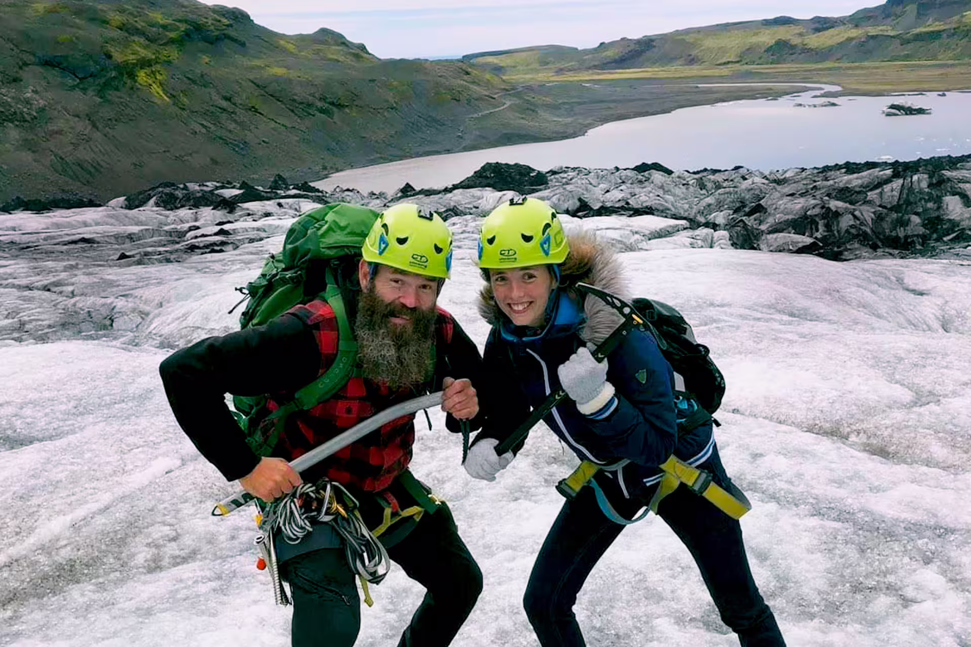 Two hikers in yellow helmets and harnesses posing on a glacier with a glacial lagoon and green mountains behind