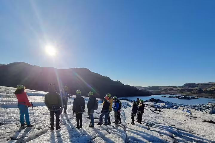 Group of hikers with helmets and trekking poles standing on a glacier with a glacial lagoon and mountains behind