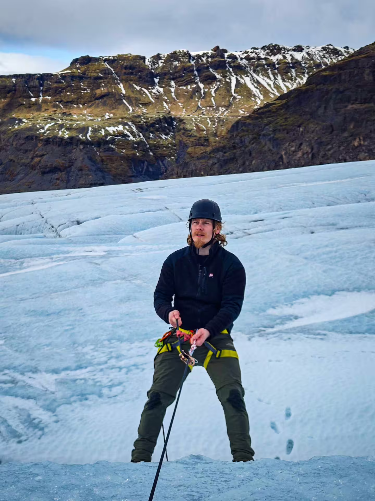 Guide in harness and helmet standing on Solheimajokull glacier with rocky mountains behind