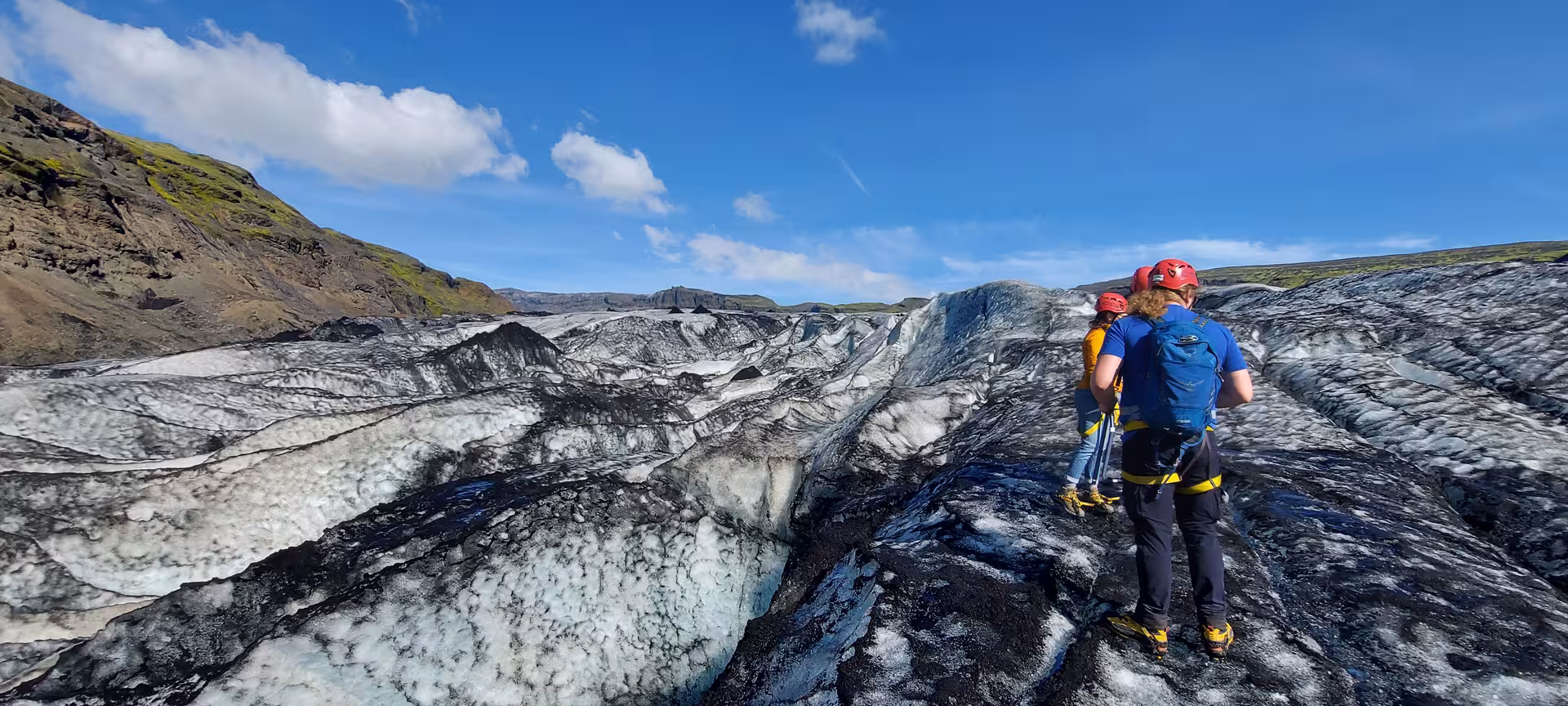 Two hikers in red helmets and crampons exploring a dark glacial ice surface in Iceland under blue skies