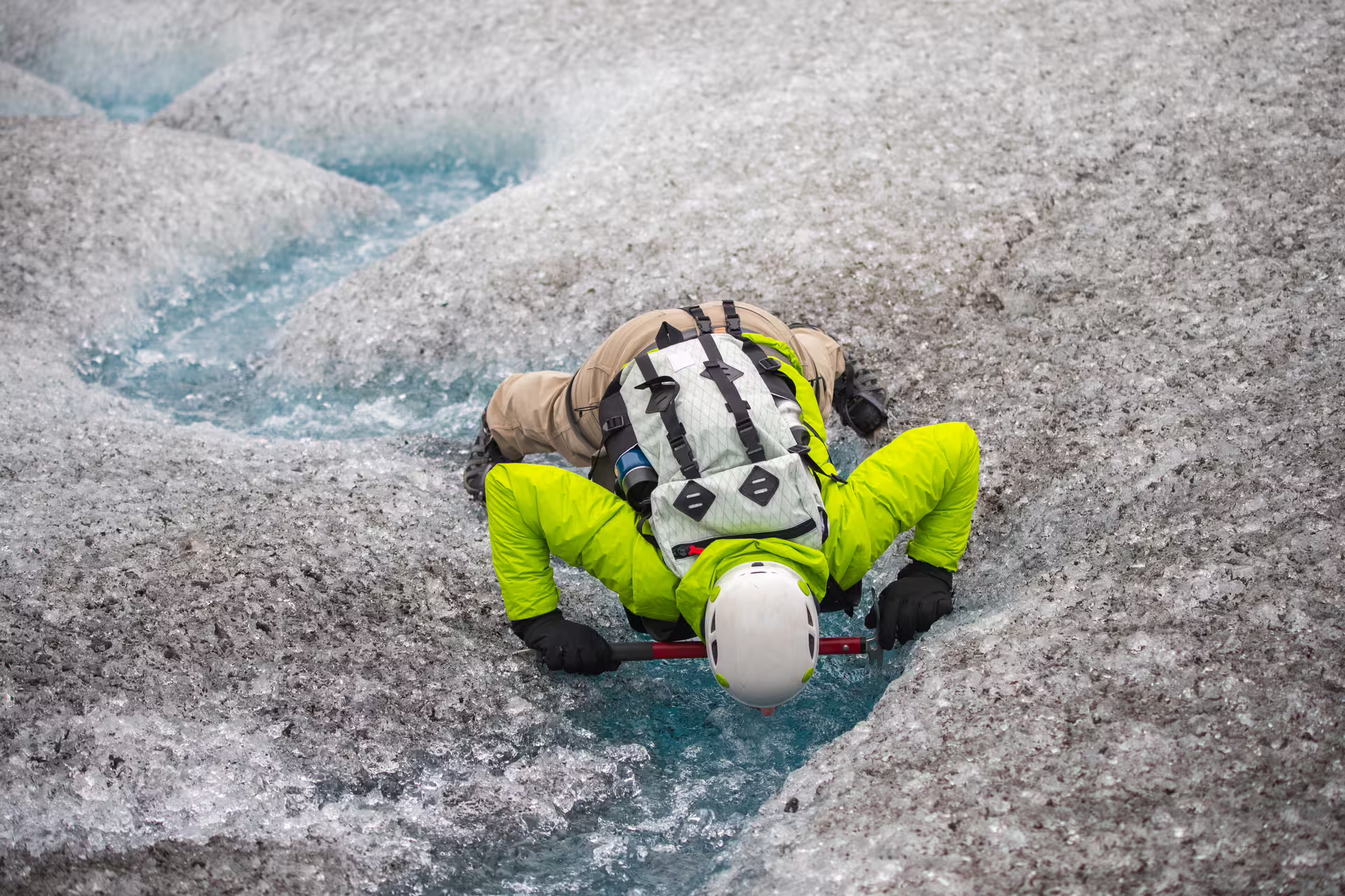 Hiker in neon yellow jacket and white helmet crossing glacial meltwater stream on Icelandic glacier