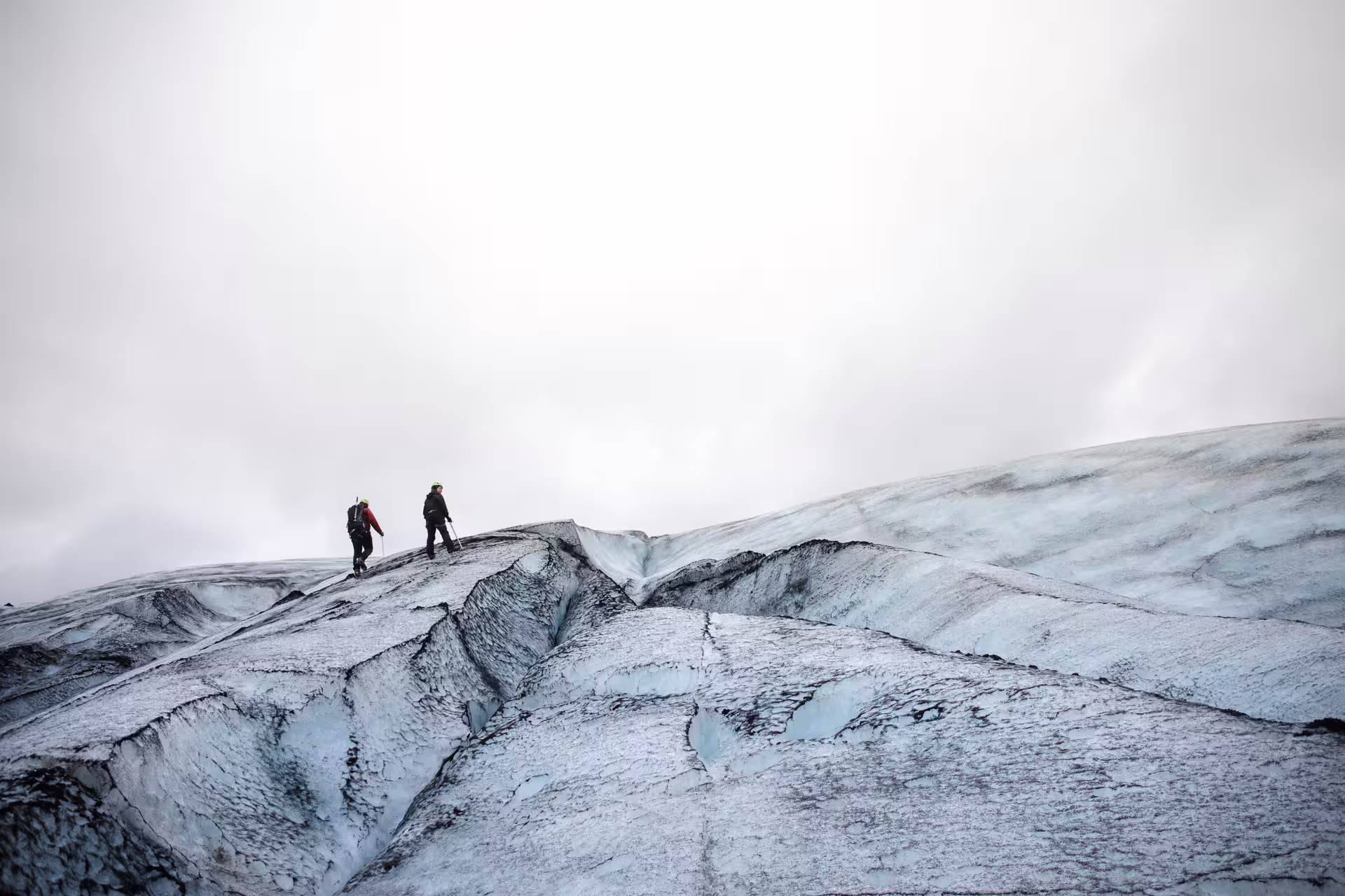Two hikers with gear walking across a cracked glacier under an overcast sky in Iceland