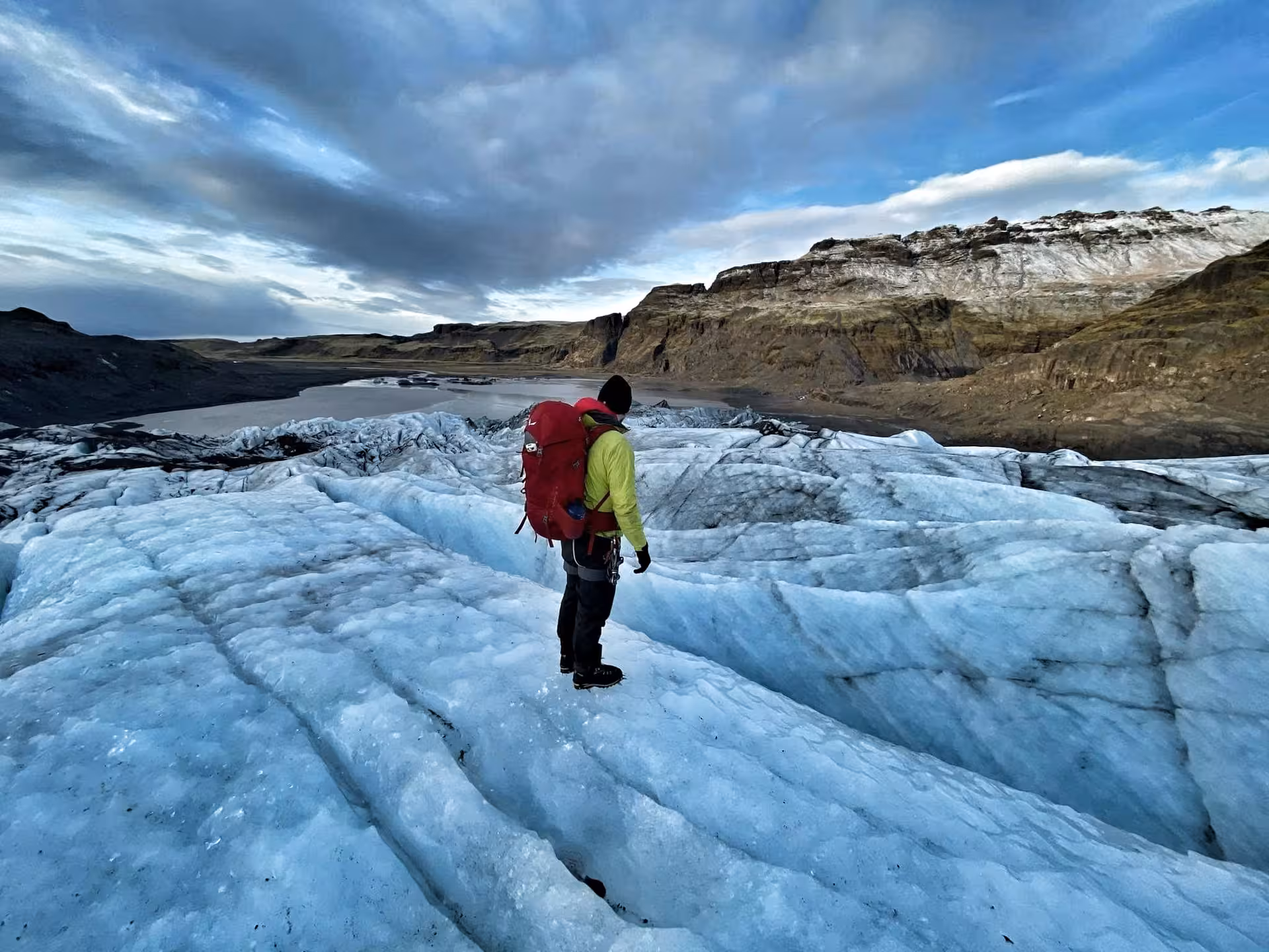Hiker with red backpack and yellow jacket standing on blue glacier ice in Iceland's south coast