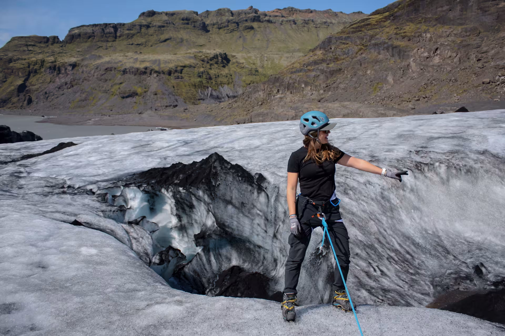 Woman in helmet and harness standing on glacier with rope near crevasse, green mountains behind
