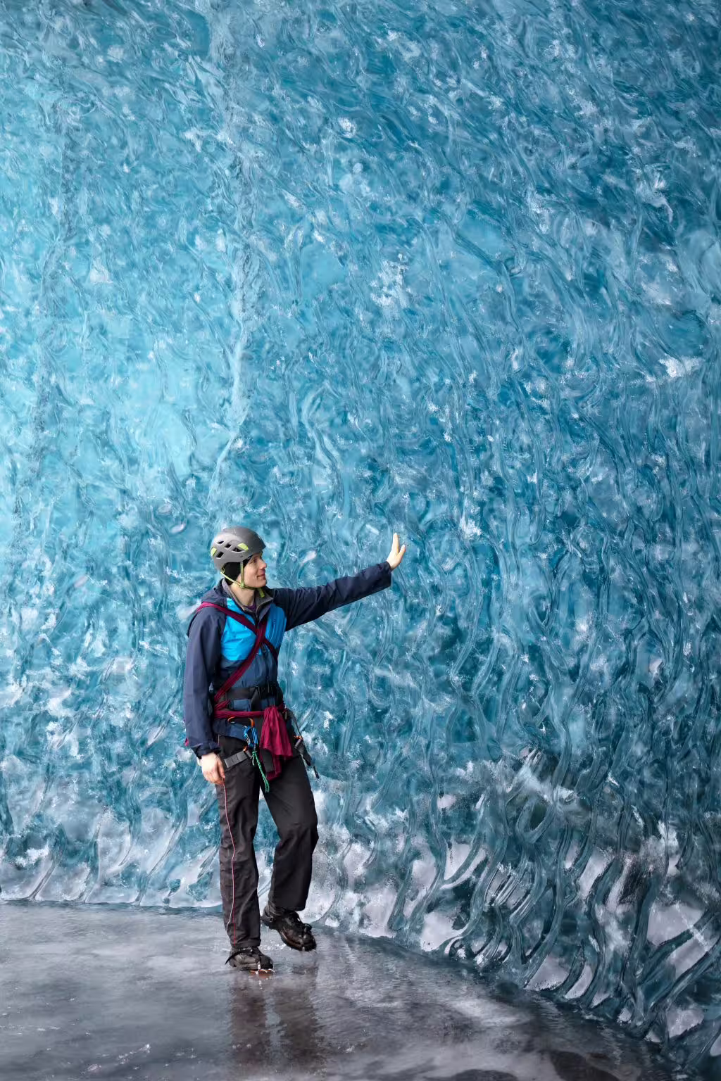 A helmeted explorer in climbing gear touches the stunning blue textured ice wall inside a glacier ice cave photoshoot adventure.