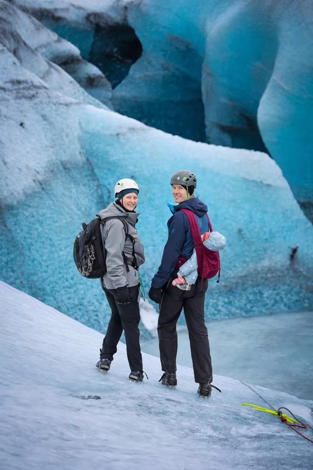 Two hikers in helmets and backpacks smiling on a glacier with stunning blue ice cave formations behind them.