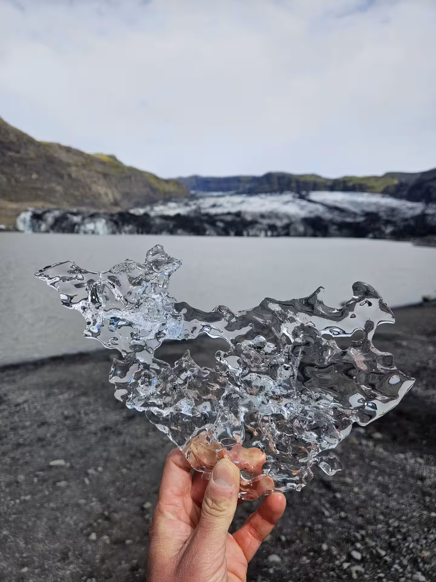 Hand holding a thin transparent glacier ice shard with glacial lagoon and ice wall in background, Iceland