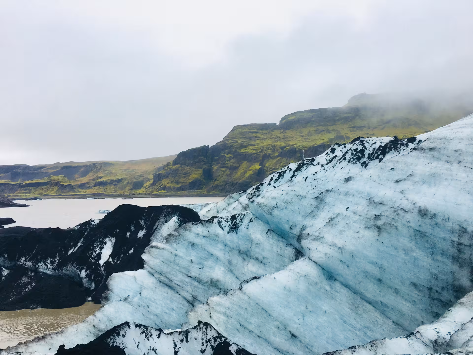 Blue glacier ice with dark volcanic rock beside a glacial lagoon and green moss-covered cliffs in Iceland