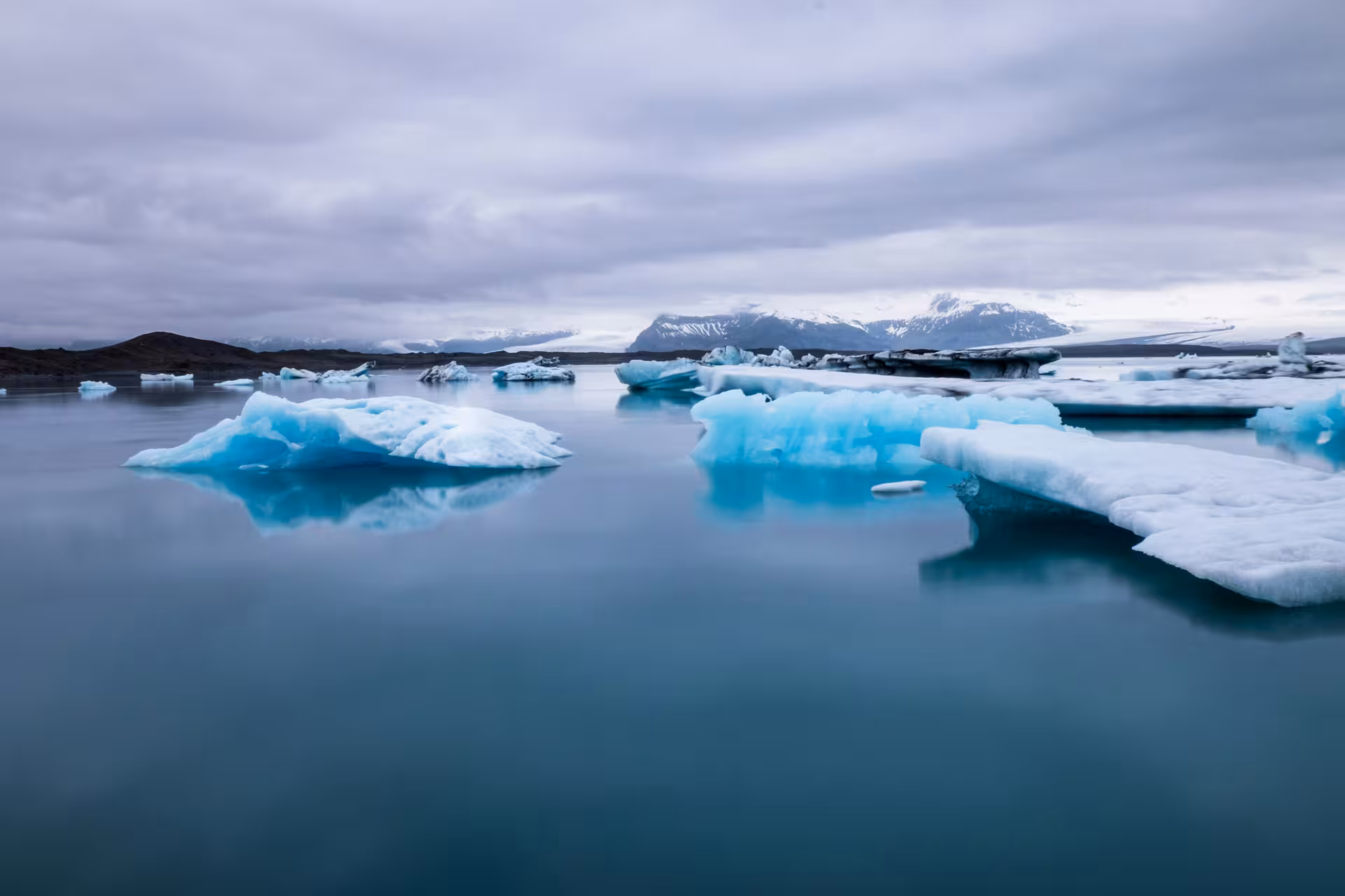 Blue icebergs floating on calm glacial lagoon waters with snow-capped mountains in background, Iceland