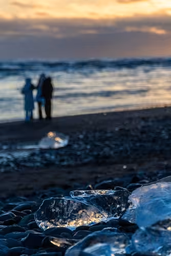 Ice chunks on black sand beach at sunset with people standing by the ocean at Glacier Lagoon, Iceland