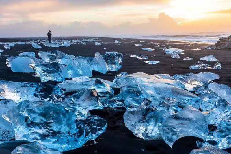 Blue ice chunks on black sand beach at Glacier Lagoon Iceland with person silhouetted at sunset