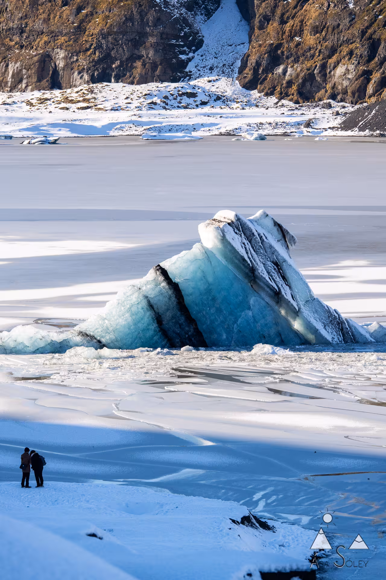 Two visitors standing before a massive teal iceberg on frozen Glacier Lagoon in Iceland in winter