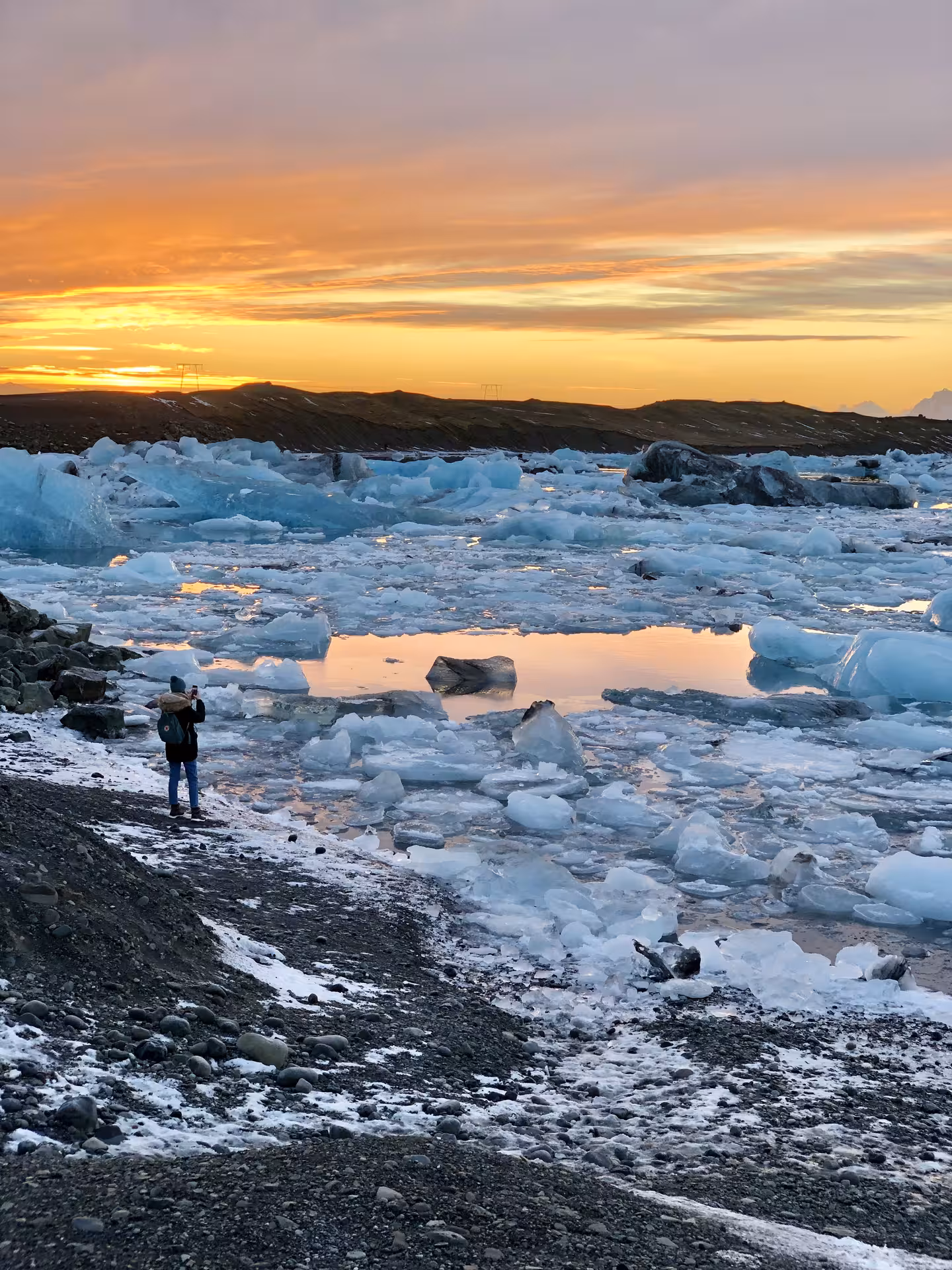 Photographer standing on black sand shore at Jokulsarlon Glacier Lagoon with blue icebergs at sunset